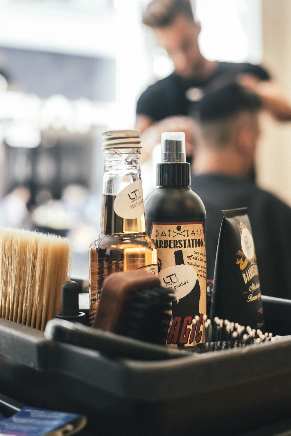 Přípravky a kartáče v boxu na barbershopovém stolku, v pozadí holič při práci.